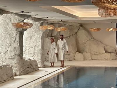 Innenraum mit großem Pool, felsähnlichen Wänden, warmem Licht und zwei Personen in Bademänteln.