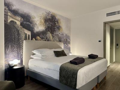 Schlafzimmer mit großem Bett, grauem Überwurf, schwarzem Nachttisch und Wandbild mit Palmen.