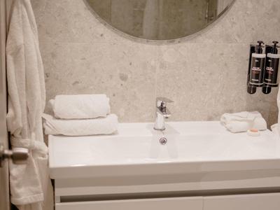 Weißes Waschbecken mit Handtüchern, runder Spiegel und Wandspendern in schlichtem Badezimmer.