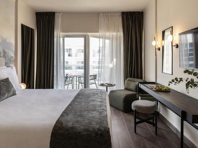Modernes Hotelzimmer mit großem Bett, Schreibtisch, Stuhl und Balkon mit Blick nach draußen.
