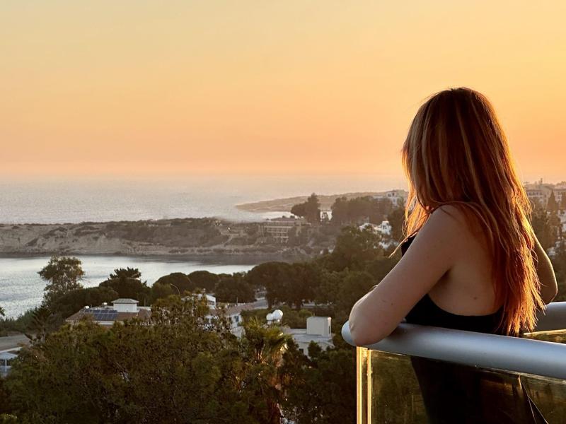 Frau mit roten Haaren blickt zum Sonnenuntergang über Küstenlandschaft und Häuser.