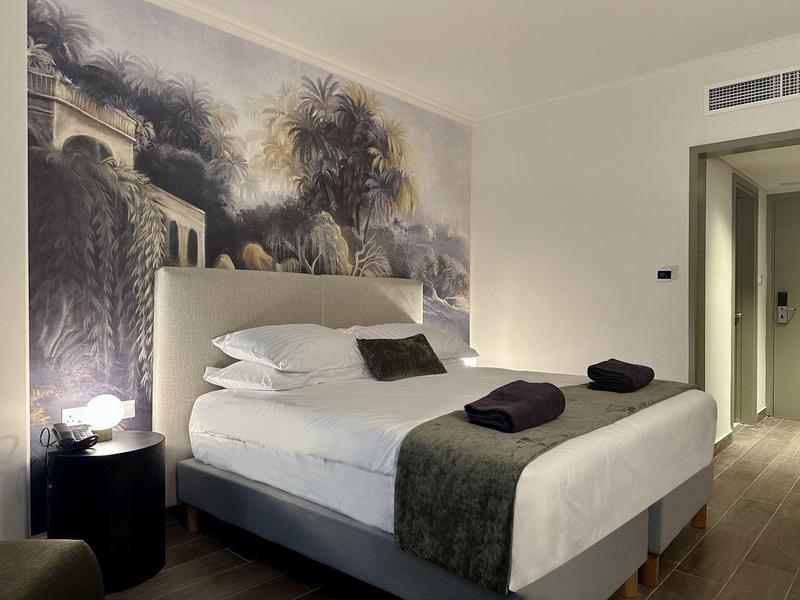 Schlafzimmer mit großem Bett, grauem Überwurf, schwarzem Nachttisch und Wandbild mit Palmen.