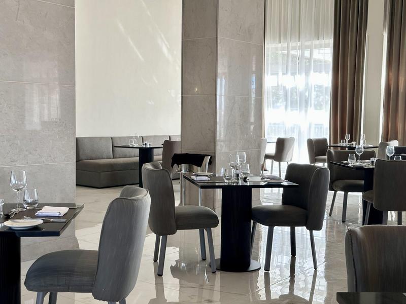 Moderner Speiseraum mit grauen Stühlen, schwarzen Tischen und großen Fenstern mit weißen Vorhängen.