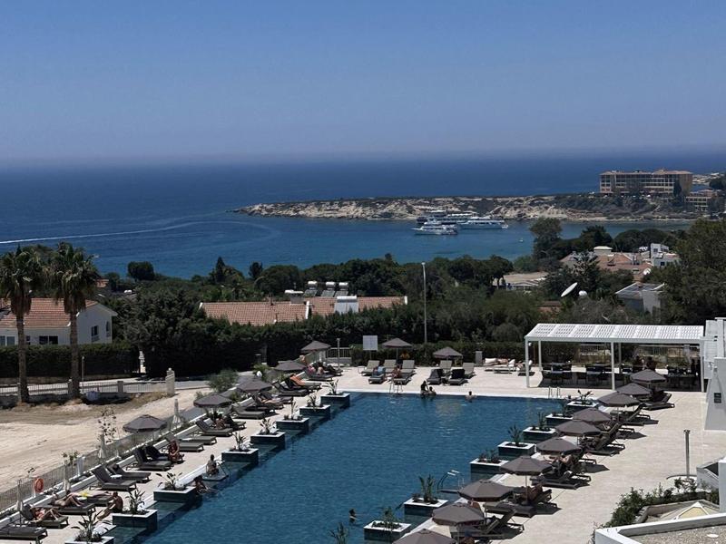 Großer Pool mit Liegen und Sonnenschirmen, dahinter Palmen und blaues Meer am klaren Himmel.