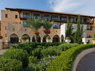 Pohled na hotelovou budovu s terasou, palmami a zelenými keři v popředí.