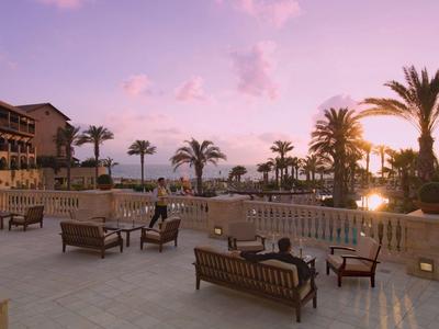 Große Terrasse mit Holzbänken, Palmen und Gebäuden bei Sonnenuntergang am Meer.