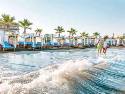 Strand mit Palmen, weißen Liegen und zwei Menschen, die durch seichtes Wasser gehen.