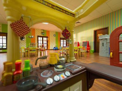 Buntes Kinderzimmer mit Spielküche, Stühlen und Tischen, hellen Wänden und Holzfenstern.