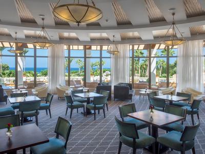 Großer, heller Speisesaal mit vielen Tischen und Stühlen, großen Fenstern und Blick auf Palmen und Meer.