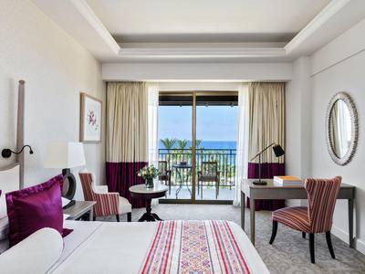 Helles Hotelzimmer mit Balkon, Meerblick, großem Bett, violetten Kissen und elegantem Schreibtisch.