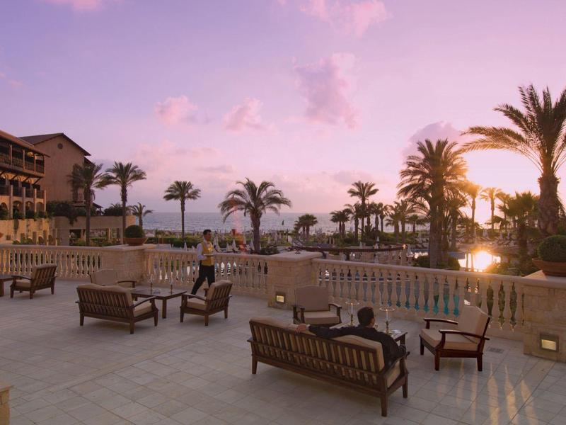 Große Terrasse mit Holzbänken, Palmen und Gebäuden bei Sonnenuntergang am Meer.