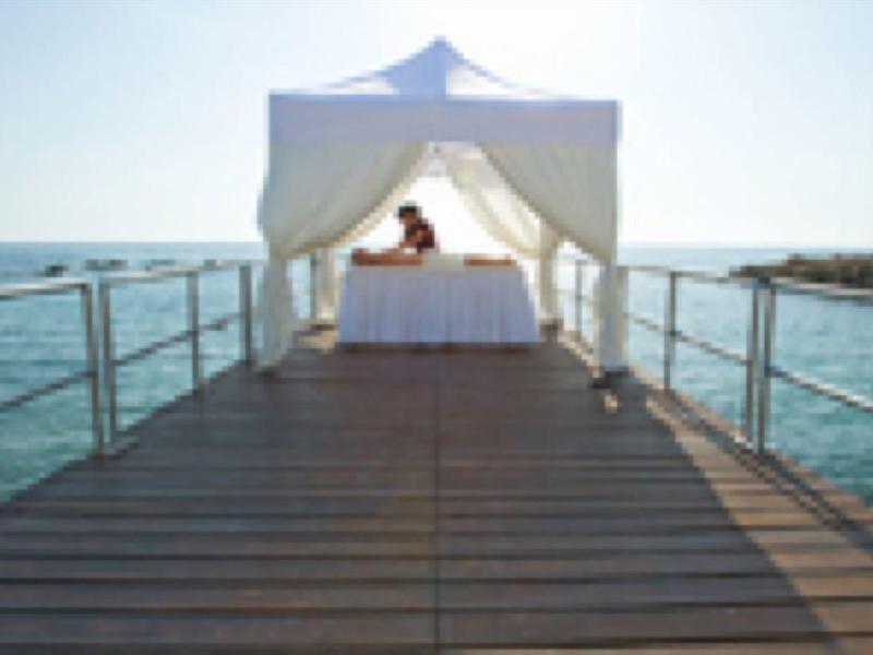 Ein Steg mit hölzernem Boden führt zu einem weißen Pavillon über dem blauen Meer.