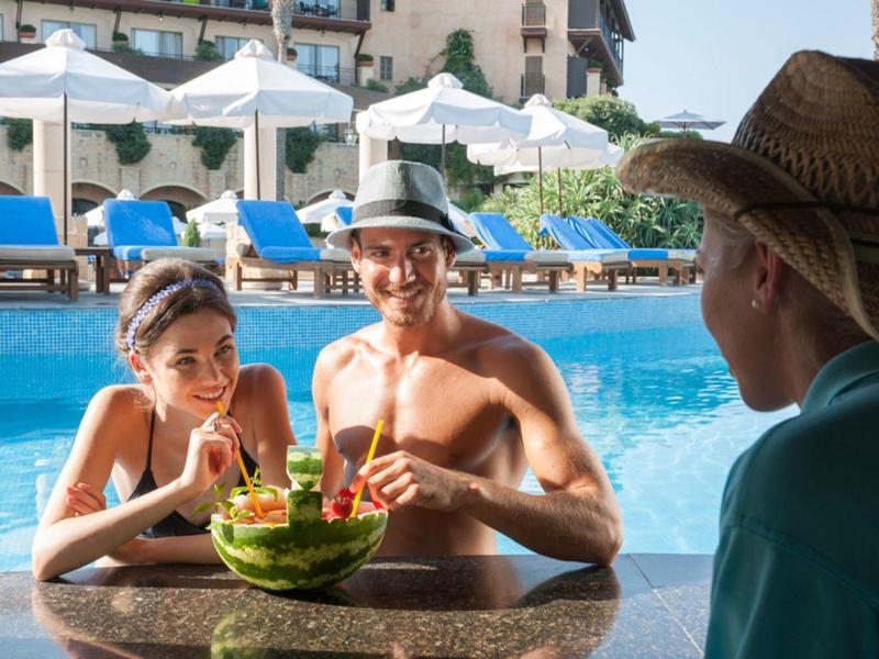 Drei Personen an Poolbar, zwei trinken aus Melonenschalen, helle Sonnenschirme und Liegestühle im Hintergrund.