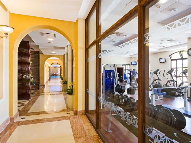 Flur mit gelben Wänden neben Fitnessraum mit Geräten und Hanteln hinter Glasscheiben.