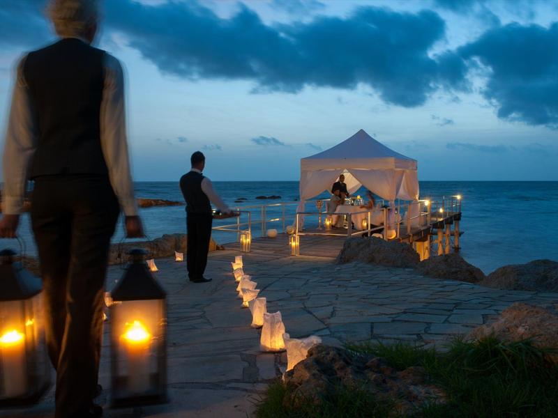 Abendliche Strandszene mit Kerzenlichtern, Personen tragen Laternen und ein Pavillon im Hintergrund.
