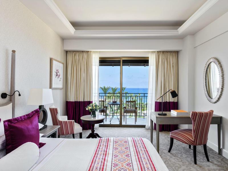 Helles Hotelzimmer mit Balkon, Meerblick, großem Bett, violetten Kissen und elegantem Schreibtisch.