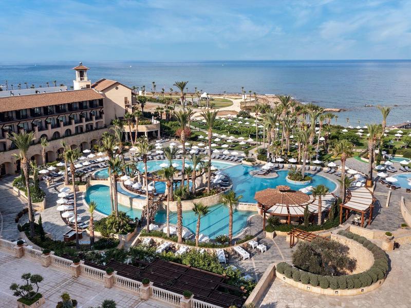 Großer Resort-Pool mit Palmen und Liegestühlen neben einem Hotel am Meer unter blauem Himmel.