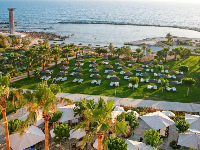 Grüne Parkanlage mit weißen Sitzgruppen, Palmen und dahinter sichtbarem Meer mit Wellenbrecher.