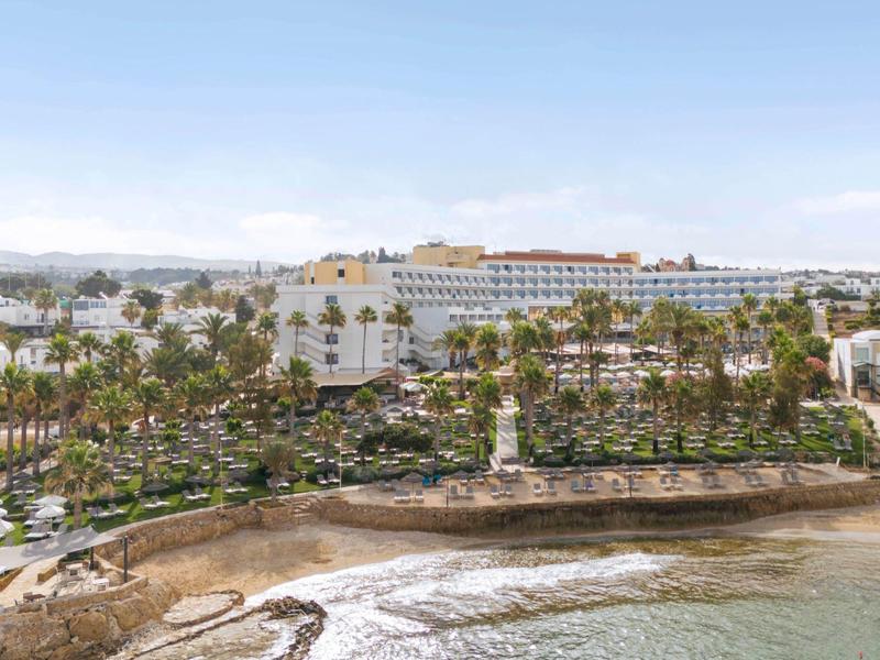 Großer weißer Hotelkomplex am Meer mit Palmen und sanften Wellen an der Küste.