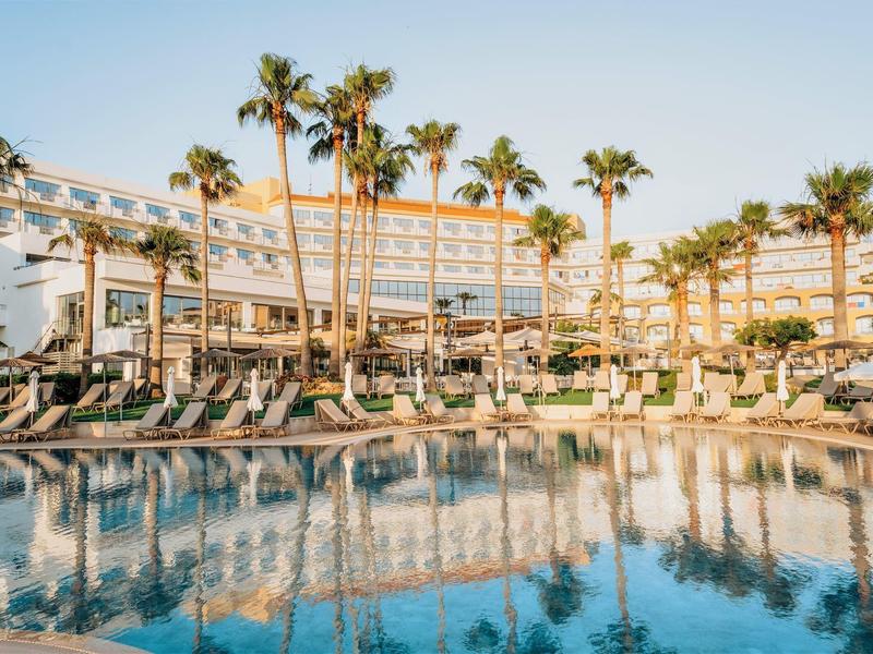Großer Pool mit Liegen und Palmen vor mehrstöckigem Hotel im warmen Sonnenlicht.