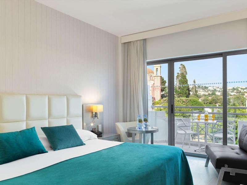 Helles Schlafzimmer mit großem Bett, türkisfarbener Decke, zwei Kissen und Balkon mit Blick nach draußen.