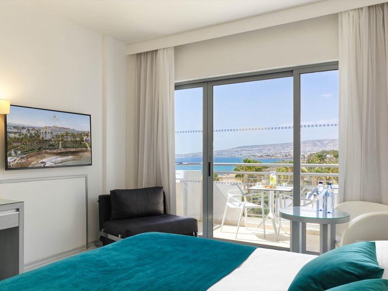 Helles Hotelzimmer mit großem Bett, Sofa, Schreibtisch, Balkon und Meerblick.