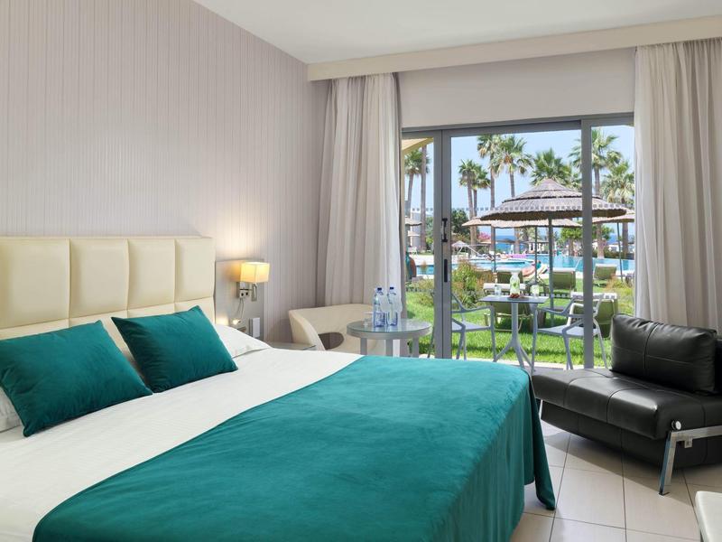 Helles Hotelzimmer mit großem Bett, türkisfarbenen Decken und Kissen, dunkelgrauem Sessel und Gartenausblick.