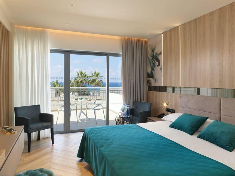 Modernes Hotelzimmer mit Doppelbett, blauen Bettdecken, schwarzem Stuhl und Balkon mit Meeresblick.