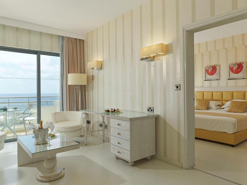 Helles Wohnzimmer mit beigen Wänden, weißem Mobiliar und Blick auf Balkon mit Meer. Schlafzimmer sichtbar.