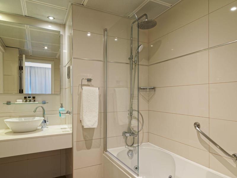 Modernes Badezimmer mit weißer Badewanne, Duschkopf, großem Spiegel und Waschbecken auf beige Fliesen.