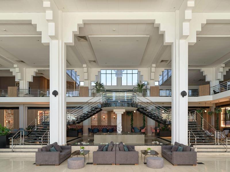 Große helle Halle mit weißen Säulen, zwei Treppenaufgängen, Sofas und großen Fenstern im Hintergrund.