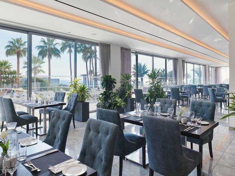 Modernes, helles Restaurant mit großen Fenstern, eleganten Stühlen und Blick auf Palmen.