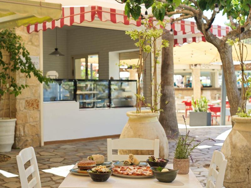 Gemütliches Café mit weißen Tischen, Stühlen, großen Töpfen und einer Auslage unter Markisen.