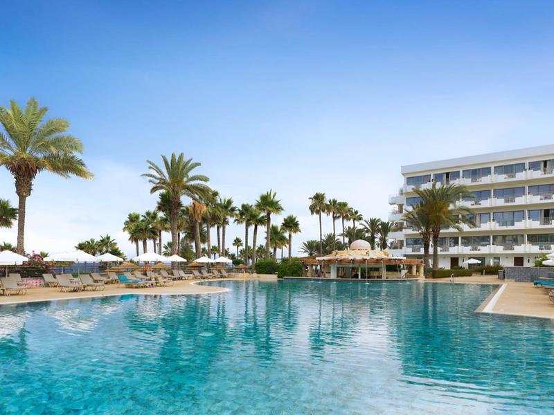 Großer blauer Pool mit Palmen und Sonnenschirmen vor einem weißen Hotelgebäude unter klarem Himmel.