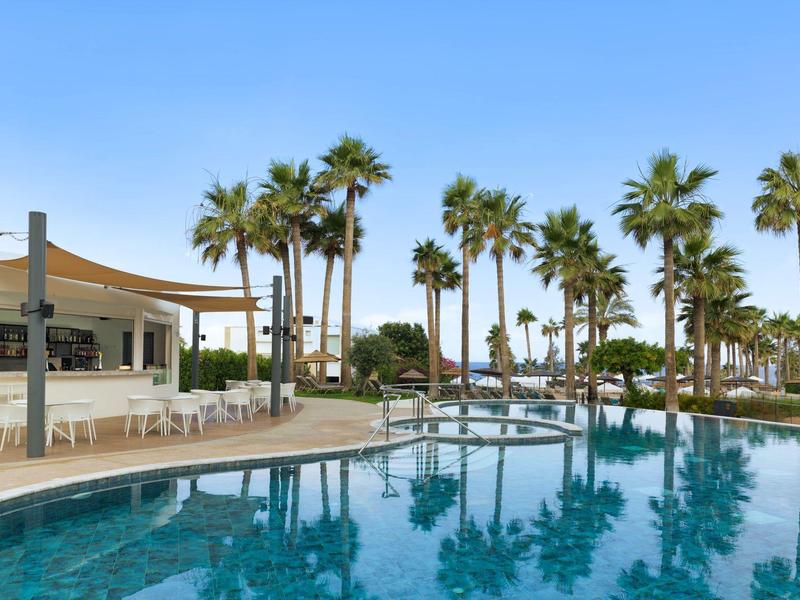 Großer Pool mit klarem Wasser, umgeben von Palmen und einem weißen Sonnensegel bei blauem Himmel.