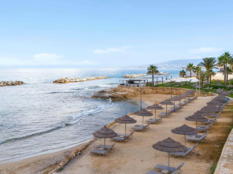 Strand mit Sand, Sonnenliegen und Strohmattenschirmen, ruhiges Meer und Palmen am Ufer.