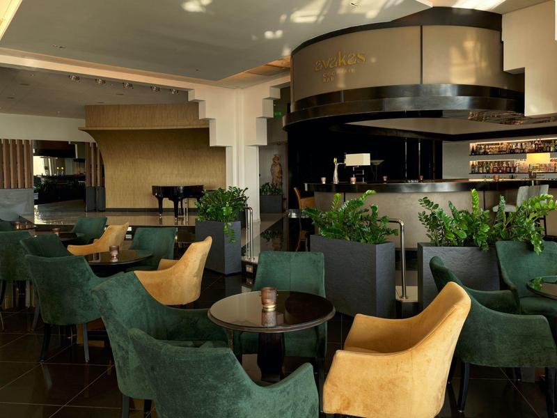 Modernes Restaurant mit grünen und gelben Sesseln, schwarzen Tischen und einer Bar im Hintergrund.