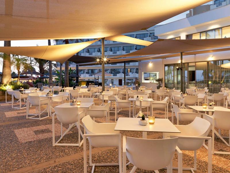 Moderner, heller Außenbereich eines Restaurants mit weißen Stühlen, Tischen und Sonnensegeln am Abend.