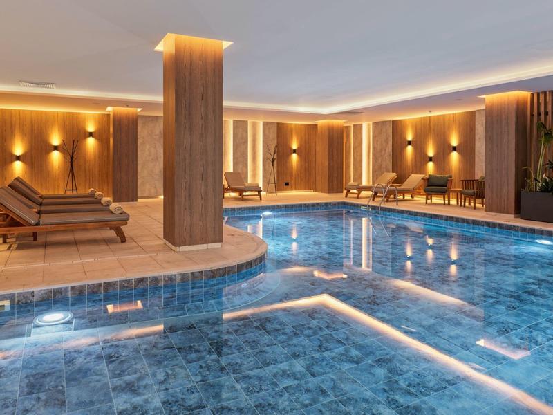 Hallenbad mit blau gefliestem Becken, beleuchteten Wänden und Holzliegen mit warmem Licht.