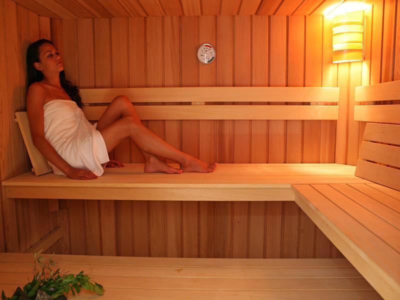 Frau entspannt in hölzerner Sauna, warmes Licht, Holzbänke und Wanduhr sichtbar.