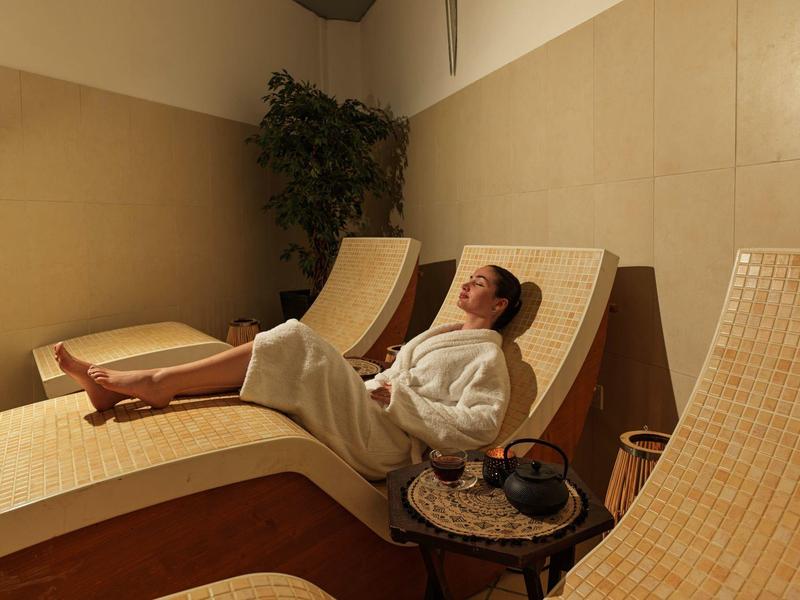 Frau entspannt im weißen Bademantel auf hellem Liegestuhl in ruhigem, warm beleuchtetem Raum.