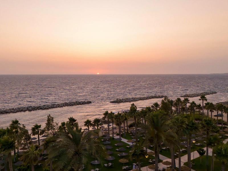 Sonnenuntergang über ruhigem Meer mit Palmen und Strand im Vordergrund.