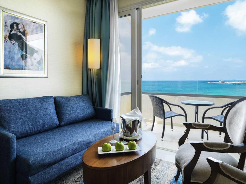 Moderner Wohnbereich mit blauem Sofa, rundem Holztisch, Getränkekühler und Meerblick vom Balkon.