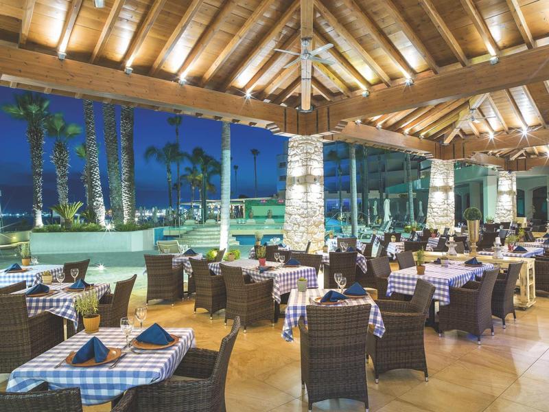 Offenes Restaurant mit gedeckten Tischen, Holzdach und Blick auf Palmen und Meer bei Abendlicht.