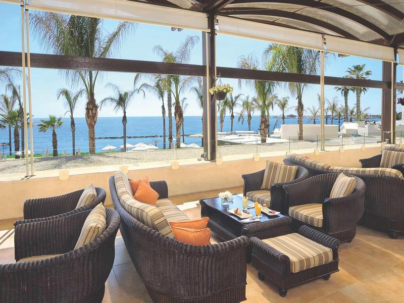 Offener Strandkorbbereich mit Sesseln und Tischen, Palmen und Meer im Hintergrund.