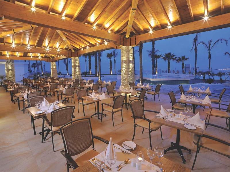 Restaurant mit gedeckten Tischen, Holzdecke, Stühlen und Blick auf Palmen und Meer bei Dämmerung.