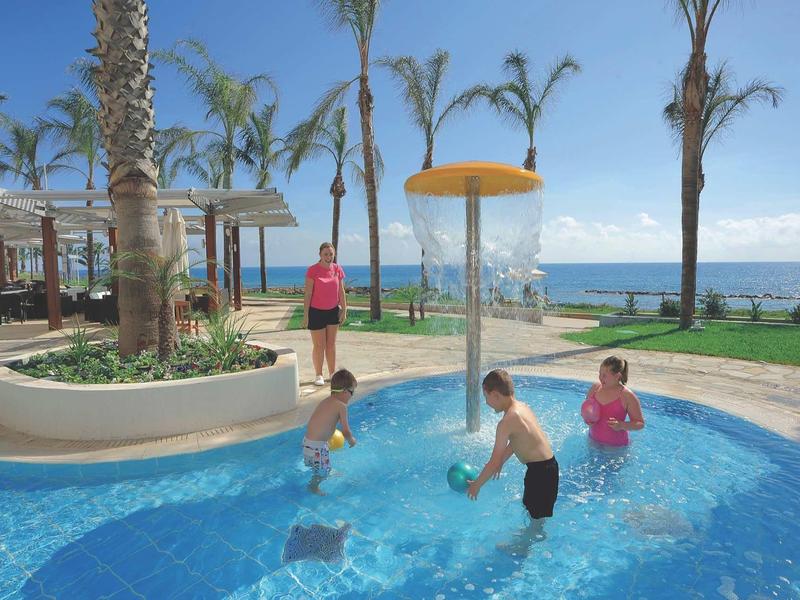 Kinder spielen mit Bällen im flachen Pool vor Palmen und Meer bei sonnigem Himmel.