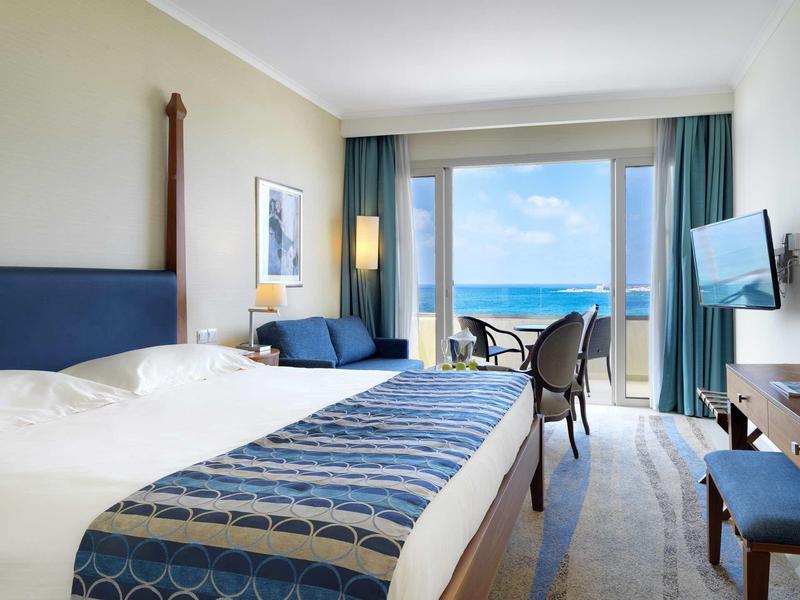 Helles Hotelzimmer mit großem Bett, blauem Sofa, Fernseher und Meerblick durch große Fenster.