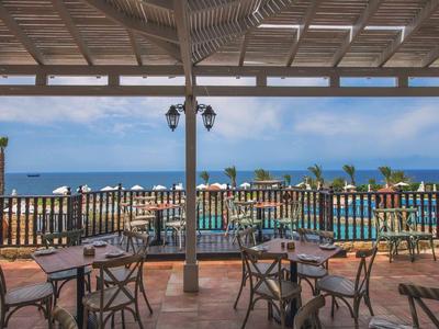 Terrasse mit Tischen und Stühlen, Blick auf Pool, Meer und blauen Himmel unter Holzdach.