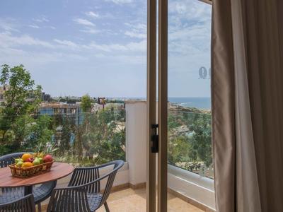 Balkon mit Tisch und Stühlen, Obstschale, Blick auf grüne Bäume und Meer am Horizont.